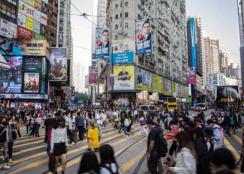 Crowds of people cross street. Tall buildings display election posters.