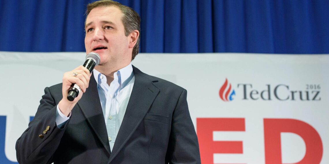 man in blazer speaks into handheld microphone in front of campaign sign
