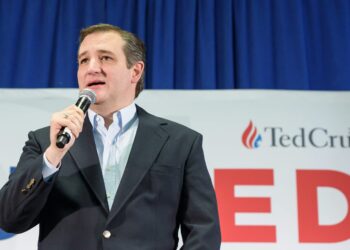 man in blazer speaks into handheld microphone in front of campaign sign