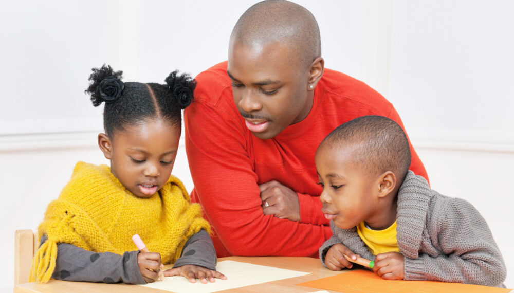 Two Black children sit at a table beside a Black man looking at worksheets. The children wear yellow attire and the man is in a red-orange shirt.