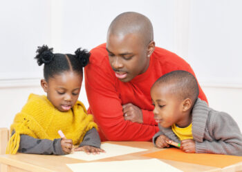 Two Black children sit at a table beside a Black man looking at worksheets. The children wear yellow attire and the man is in a red-orange shirt.