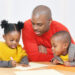 Two Black children sit at a table beside a Black man looking at worksheets. The children wear yellow attire and the man is in a red-orange shirt.