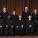 official portrait of nine justices wearing black judicial robes. five justices sit in chairs while four justices stand behind them.