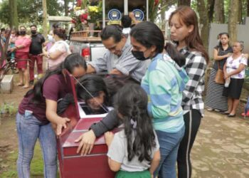 Young women and a man lean over a red coffin crying, while other people stand around solemnly
