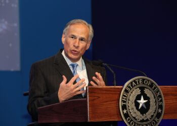 man holds both hands close to chest while speaking behind lectern bearing a texas seal