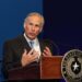 man holds both hands close to chest while speaking behind lectern bearing a texas seal