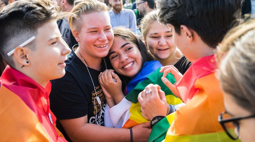 Older children smile and hug while wearing the Pride flag.