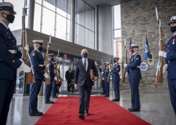 man wearing suit and face mask strides down red carpet as men in military garb stand at attention on both sides