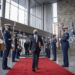 man wearing suit and face mask strides down red carpet as men in military garb stand at attention on both sides