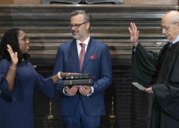 ketanji brown jackson stands with her right hand raised, stephen breyer stands facing her with his right hand raised while reading from piece of paper, jackson's husband stands between them looking at jackson