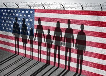 Immigrants behind a wall painted with the American flag and topped with barbed wire