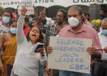 Protesters shout slogans. One has a sign saying “Release apprehended union leaders”