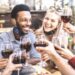Friends toasting red wine at outdoor restaurant bar with open face mask – New normal lifestyle concept with happy people having fun together on warm filter – Focus on afroamerican guy