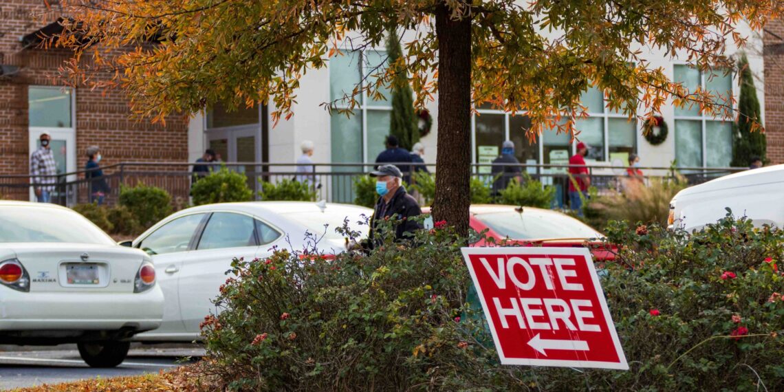 parking lot with "vote here" sign and line of people waiting outside in background