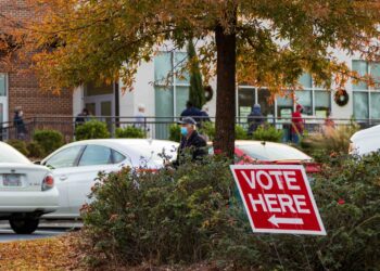 parking lot with "vote here" sign and line of people waiting outside in background