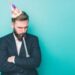 Sad guy is standing and looking down. He is upset. Man is holding a wistle in his mouth and has a birthday hat on the head. Isolated on blue background.