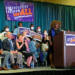 People gathered on a stage listening to a woman at a lecturn. Signs reading Reproductive Freedom for All are hung all over.