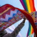 Back of person in white shirt and hat holding rainbow pride flag in the air alongside a colorful designed scarf.