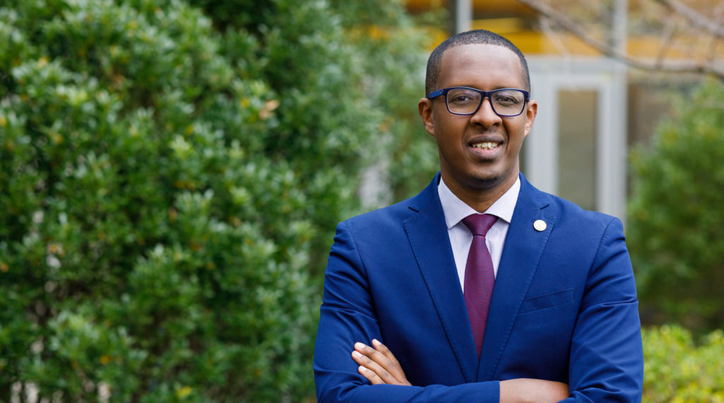 Benyam Dawit Mezmur wearing a blue suit and standing in front of trees with his arms crossed.