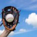Hand holding baseball in glove with blue sky