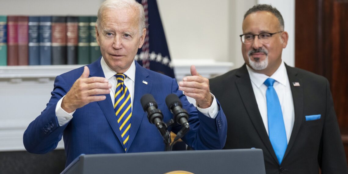 joe biden at lectern, gesturing with both hands, as miguel cardona looks on