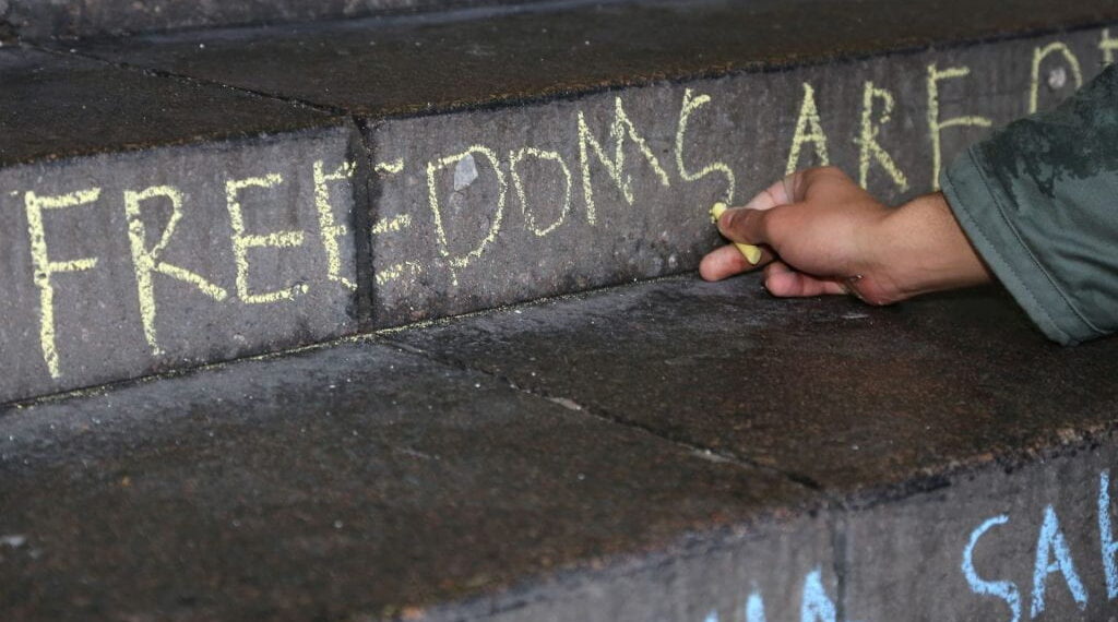 Image shows a person writing the word freedoms using chalk on a concrete staircase.