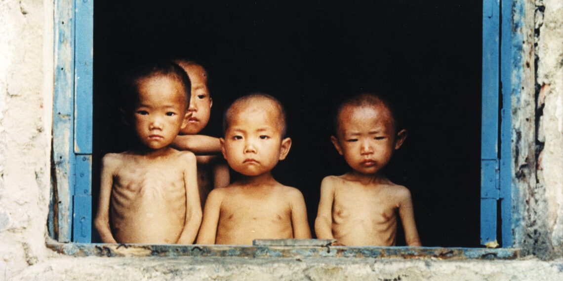 Four young Korean children stare sorrowfully through an open window with blue doors. Their ribs are visible and their arms are skinny.
