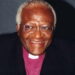 A headshot of a Black man smiling wearing purple clerical attire and collar.