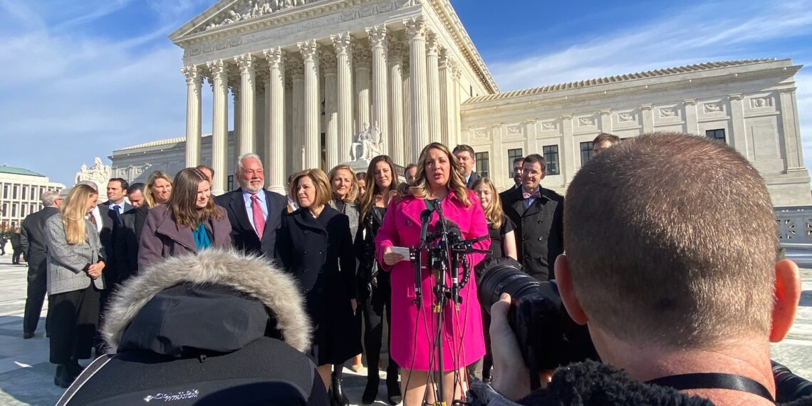 A woman in a winter coat speaks at a microphone, surrounded by supporters, with the Supreme Court building in the background.
