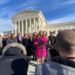 A woman in a winter coat speaks at a microphone, surrounded by supporters, with the Supreme Court building in the background.