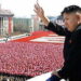 Kim Jong-Un stands on a high ledge waving to thousands of people standing uniformly down below. He is wearing a black suit. Everyone in the crowd is wearing red, and there is a group of people in the distance wearing white to spell something out among the red. It is not clear what they are positioned to spell out.