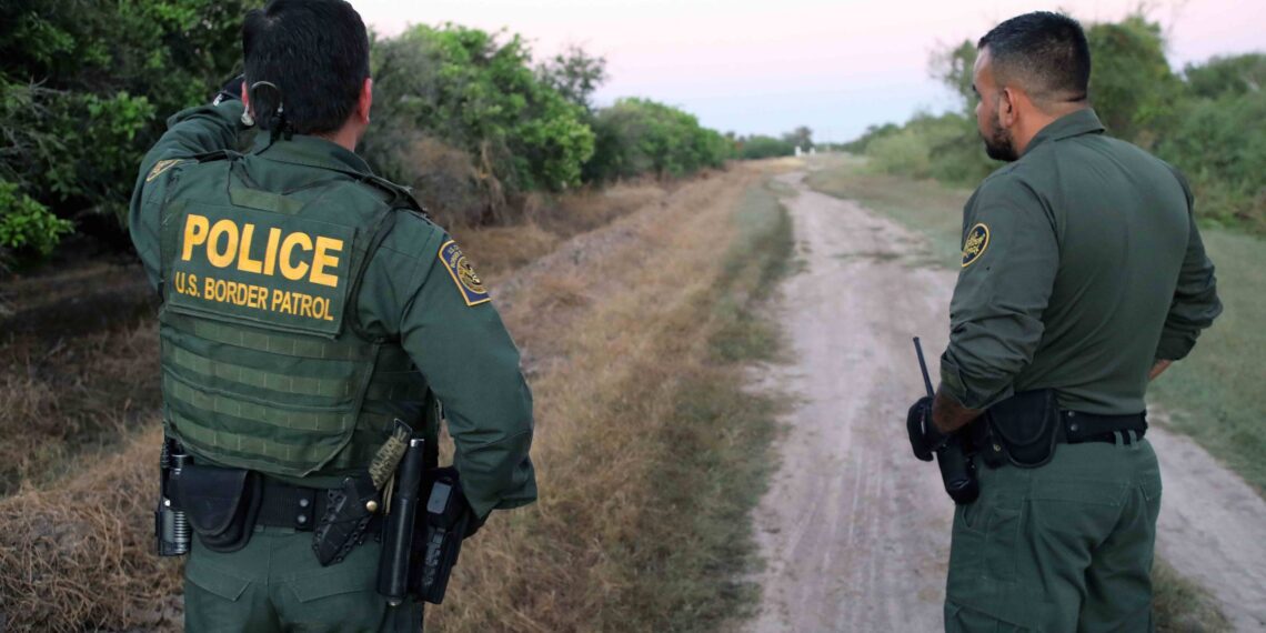 two men wearing border parol uniforms stand along dirt road with back to the camera. one points into the distance.