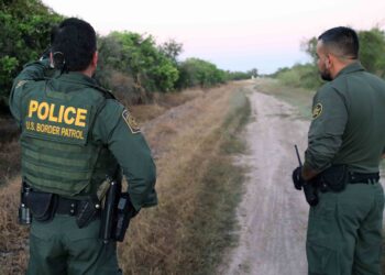 two men wearing border parol uniforms stand along dirt road with back to the camera. one points into the distance.