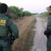 two men wearing border parol uniforms stand along dirt road with back to the camera. one points into the distance.