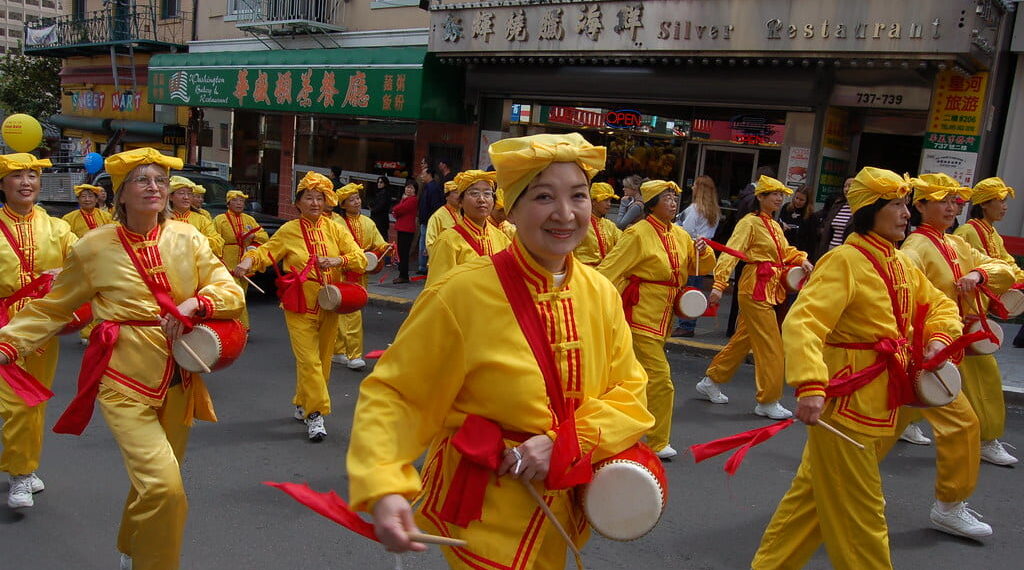A parade of people wearing bright yellow outfits with red accents walk down the streets with drums.