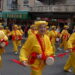 A parade of people wearing bright yellow outfits with red accents walk down the streets with drums.