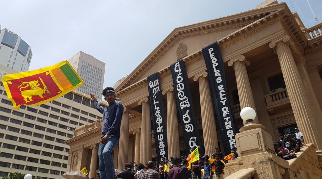 Protestor waving Sri Lankan flag in front of imposing government building.