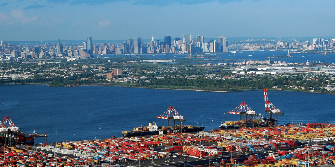 aerial view of busy seaport crowded with containers. a body of water separates the port in the foreground from the manhattan skyline in the background.