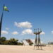 A large plaza with a statue of two elongated figures, the Brazilian flag flying, and buildings in the distance.