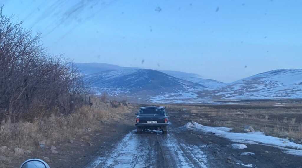 A photo of the back of an old car driving on a muddy road in front of snow-covered hills. The photo was taken from the front seat of a different car, and the hood is visible at the bottom.