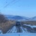 A photo of the back of an old car driving on a muddy road in front of snow-covered hills. The photo was taken from the front seat of a different car, and the hood is visible at the bottom.