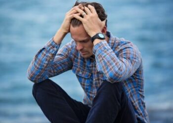 man sitting by a body of water with his head in his hands looking anxious