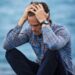 man sitting by a body of water with his head in his hands looking anxious
