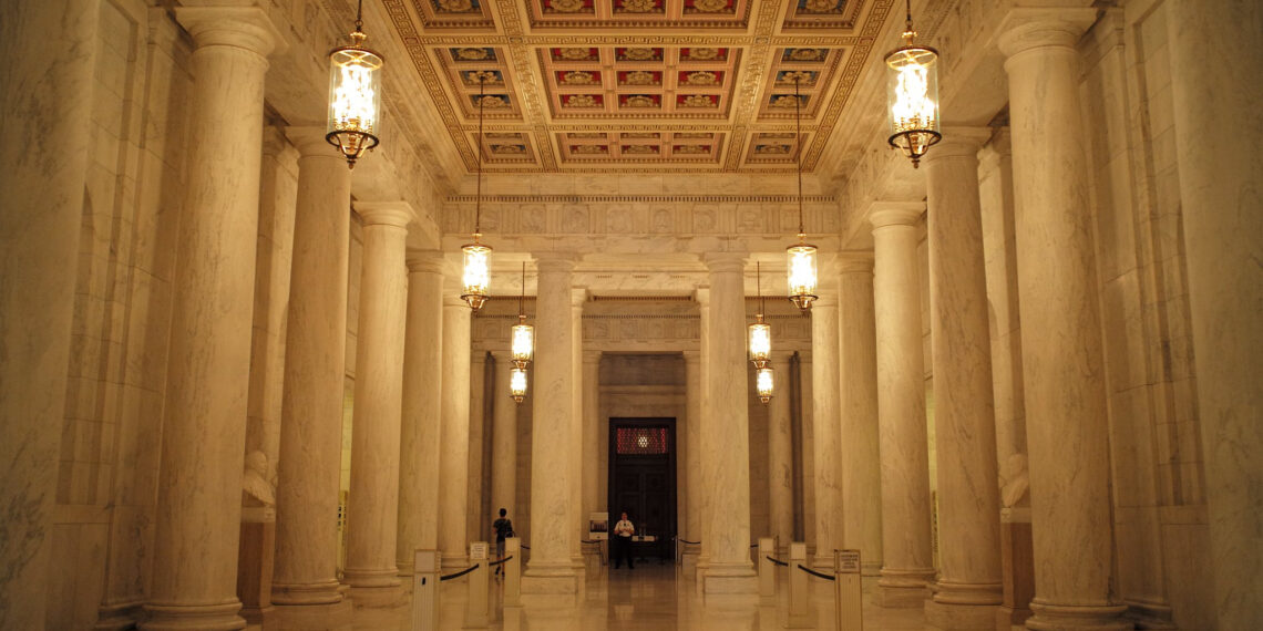 A long marble lobby with hanging lamps and marble columns. A guard waiting by a door at the end of the hall.