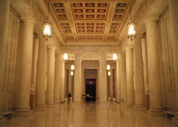 A long marble lobby with hanging lamps and marble columns. A guard waiting by a door at the end of the hall.