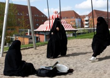 Two women on swing and one sitting in sand of play park, all dressed in black burqas