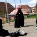 Two women on swing and one sitting in sand of play park, all dressed in black burqas