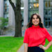 Rebecca Gore is wearing a red top and navy pants and is smiling into the camera. She is standing in front of the Harvard Law School Library.