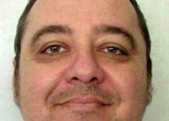 A mug shot of a smiling, middle-aged, balding white man, whose brown hair is combed back from the crown of his head. He has stubble and appears to be wearing a white-collared shirt.