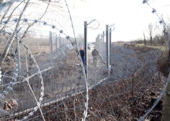 Razor wire in the foreground frames a view of the border fence between Hungary and Serbia, with two guards waling along it